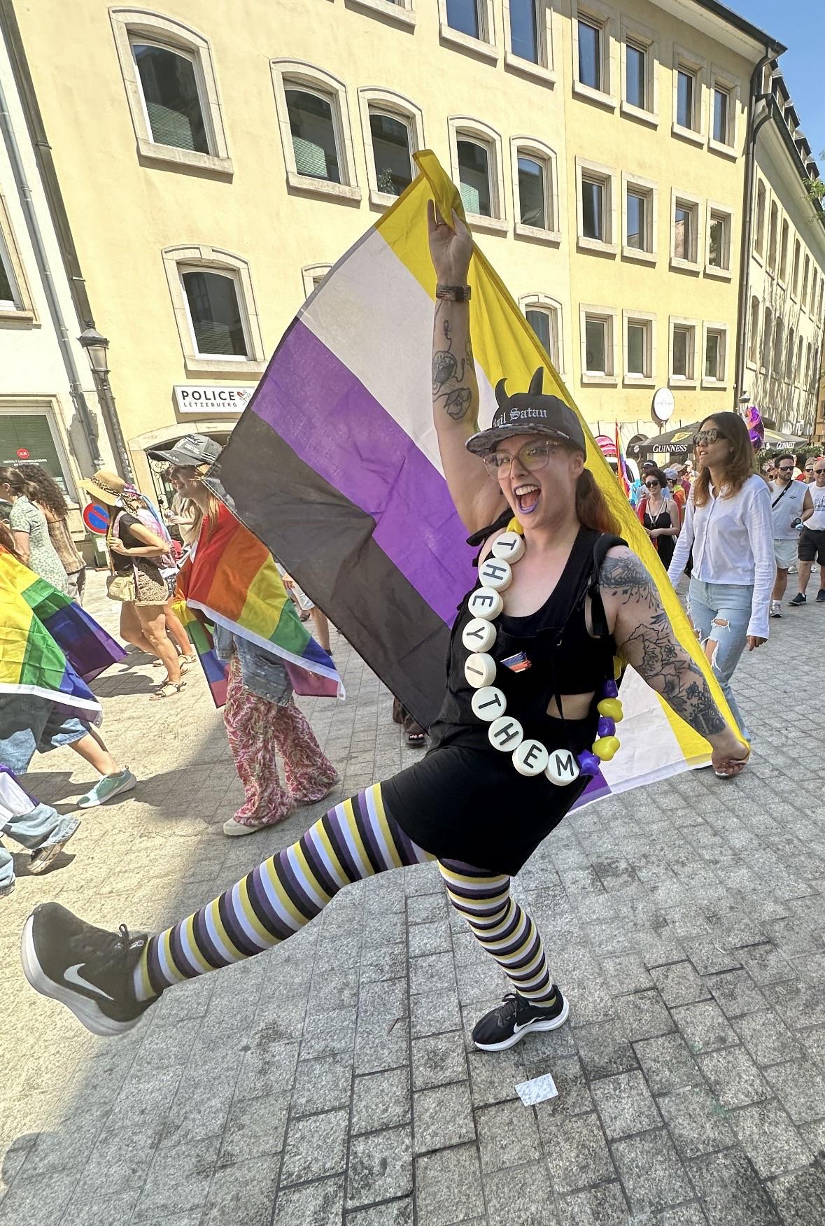 I’m posing with my non binary flag and tights, my « hail satan » hat with horns and a « they/them » giant friendship bracelet around my body